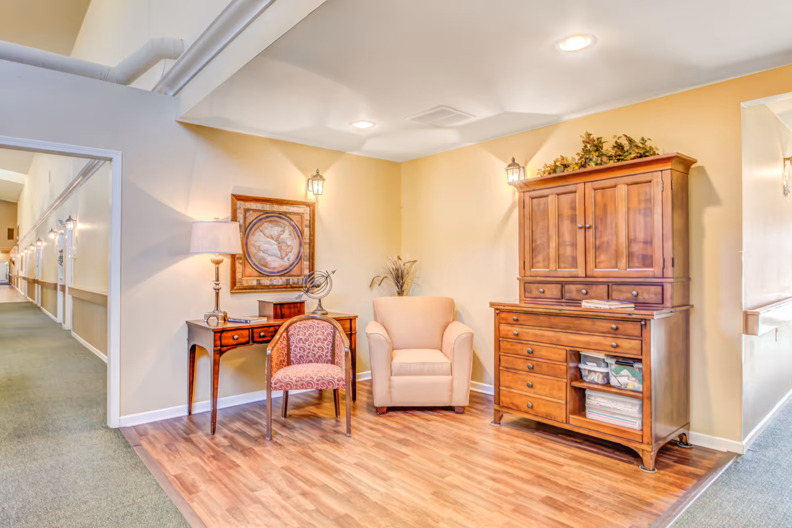 A cozy corner in an assisted living facility featuring a wooden cabinet with drawers and shelves, a beige armchair, a patterned upholstered chair, a wooden desk with a lamp and decorative items, and a framed map on the wall. The area has warm yellow walls and wood flooring, with a carpeted hallway visible to the left.