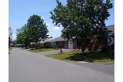 A paved road lined with single-story residential buildings on the right side, each with a small front yard and mature trees providing shade. The sky is clear and blue.