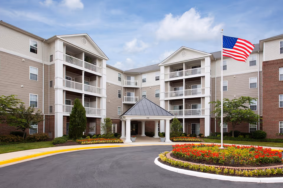 Front exterior view of a senior living facility named Manchester Lakes Senior Apartments, featuring a multi-story building with balconies, a covered entrance with white columns, landscaped flower beds, and an American flag on a flagpole.