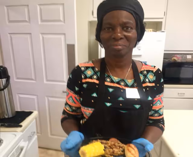 A woman wearing a colorful patterned shirt, black apron, and black head covering is standing in a kitchen holding a plate of food with corn on the cob, a bread roll, and a serving of mixed vegetables or beans. The kitchen has white cabinets, a microwave, and a refrigerator in the background.