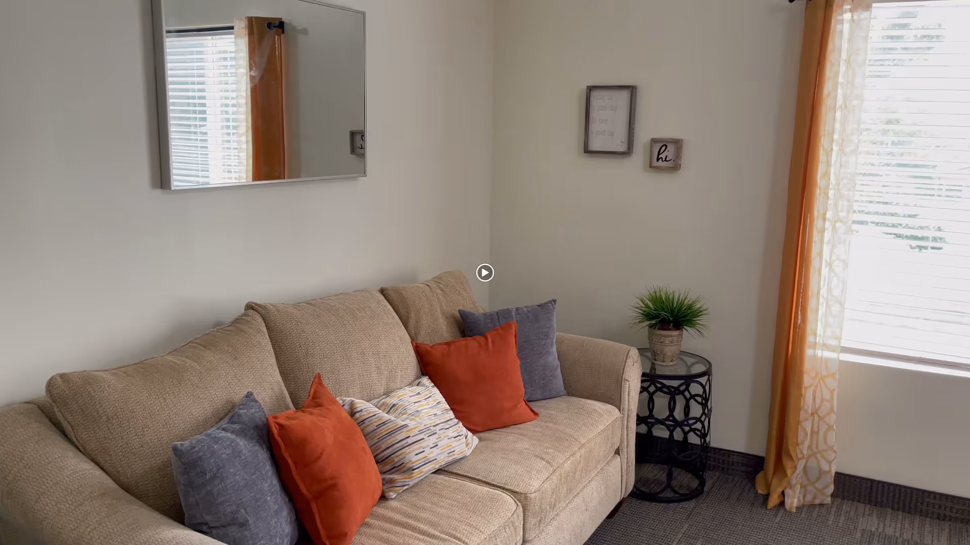 A cozy living room area with a beige couch adorned with two red, two gray, and one patterned throw pillows. A small round black side table with a potted green plant sits next to the couch. A rectangular mirror hangs on the wall above the couch, and two small framed wall decorations are on the adjacent wall. A window with white blinds and orange patterned curtains allows natural light into the room.