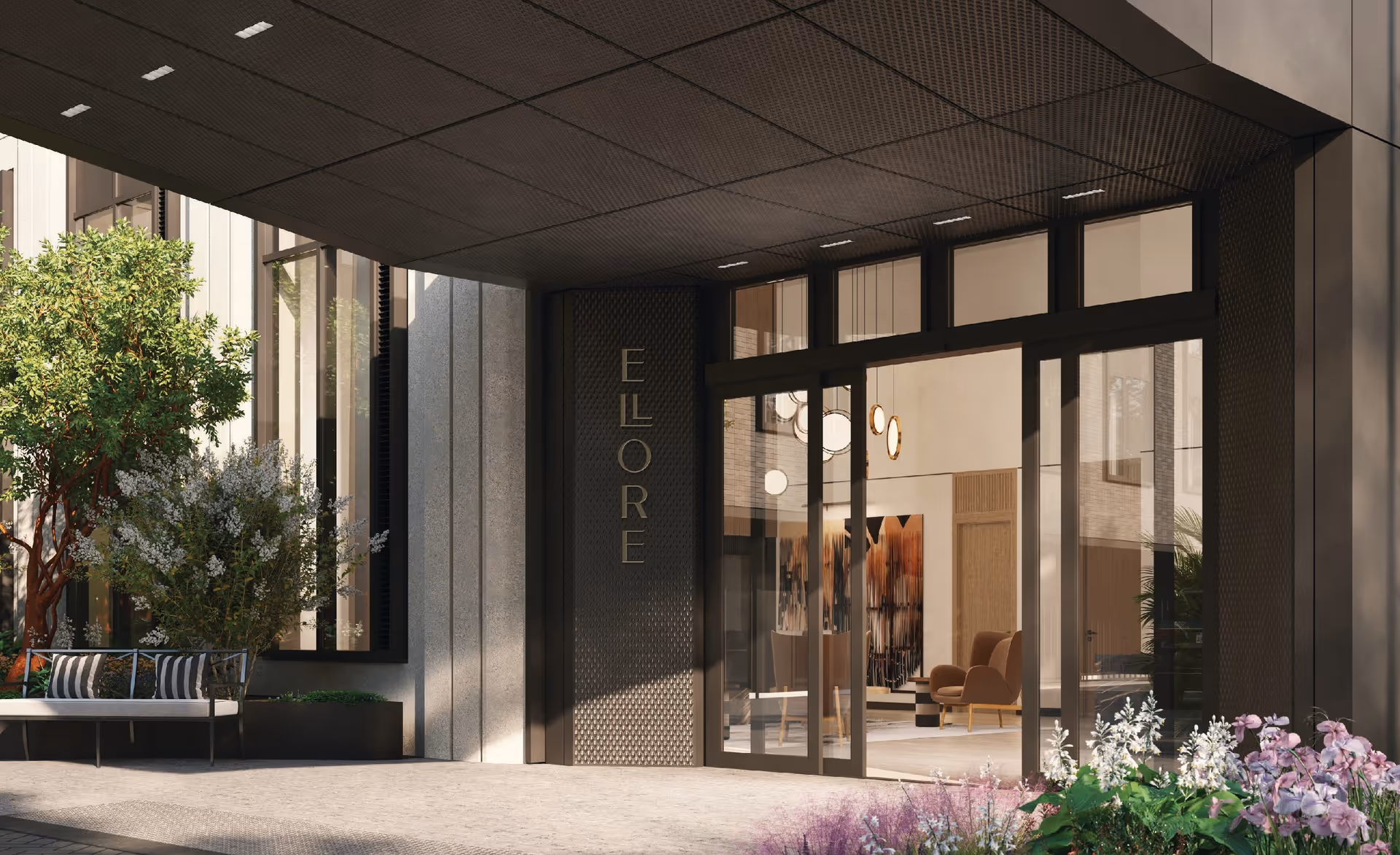 Entrance of Ellore Senior Living facility showing a modern building facade with large glass doors, a bench with cushions, greenery including trees and flowering plants, and a covered entryway with the name 'ELLORE' displayed vertically on a textured panel.