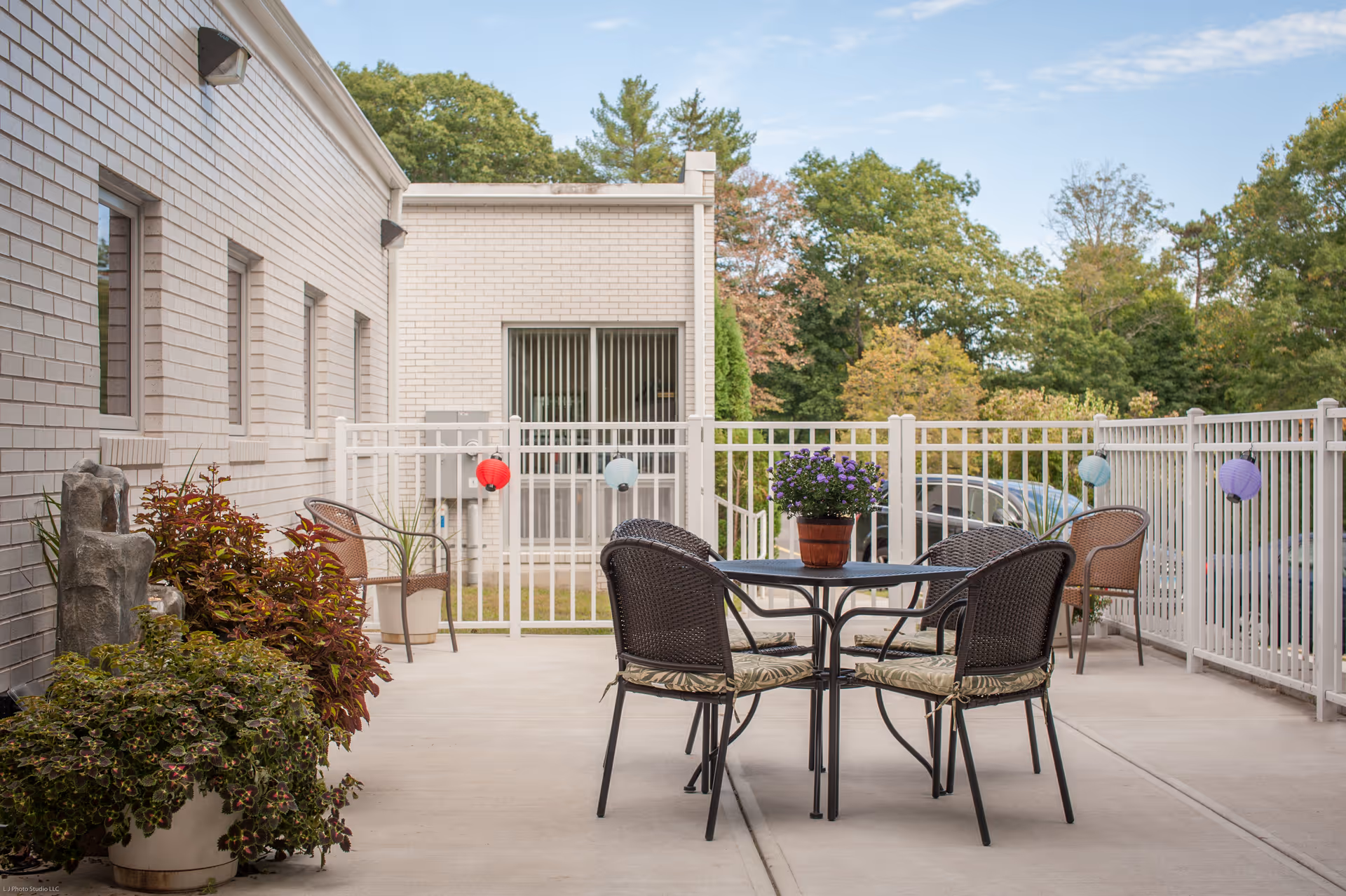 Outdoor patio area at Complete Care at Middlebury with a round table and four chairs, potted plants, and a white fence decorated with colorful lanterns. Trees and greenery are visible in the background.