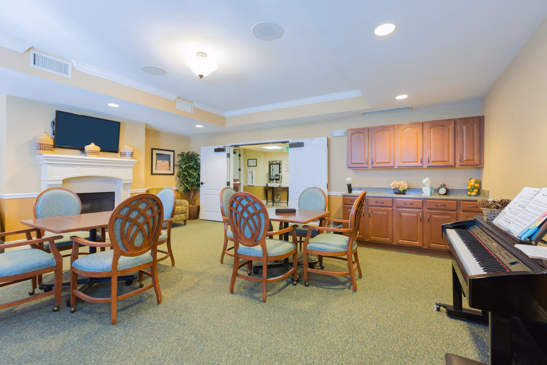 A bright common room with round tables and wooden upholstered chairs, a fireplace with TV, built-in cabinets, and a piano.