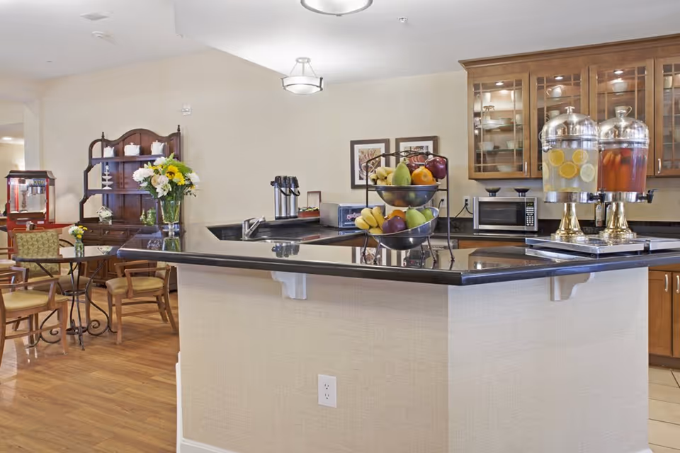 A bright and clean kitchen area in a senior living facility with a black countertop island featuring a two-tiered fruit basket filled with bananas, apples, pears, and oranges. Behind the island, there are wooden cabinets with glass doors, a microwave, and two beverage dispensers containing lemon water and a red drink. To the left, there is a small dining area with a table, chairs, a wooden hutch with decorative items, and a popcorn machine.
