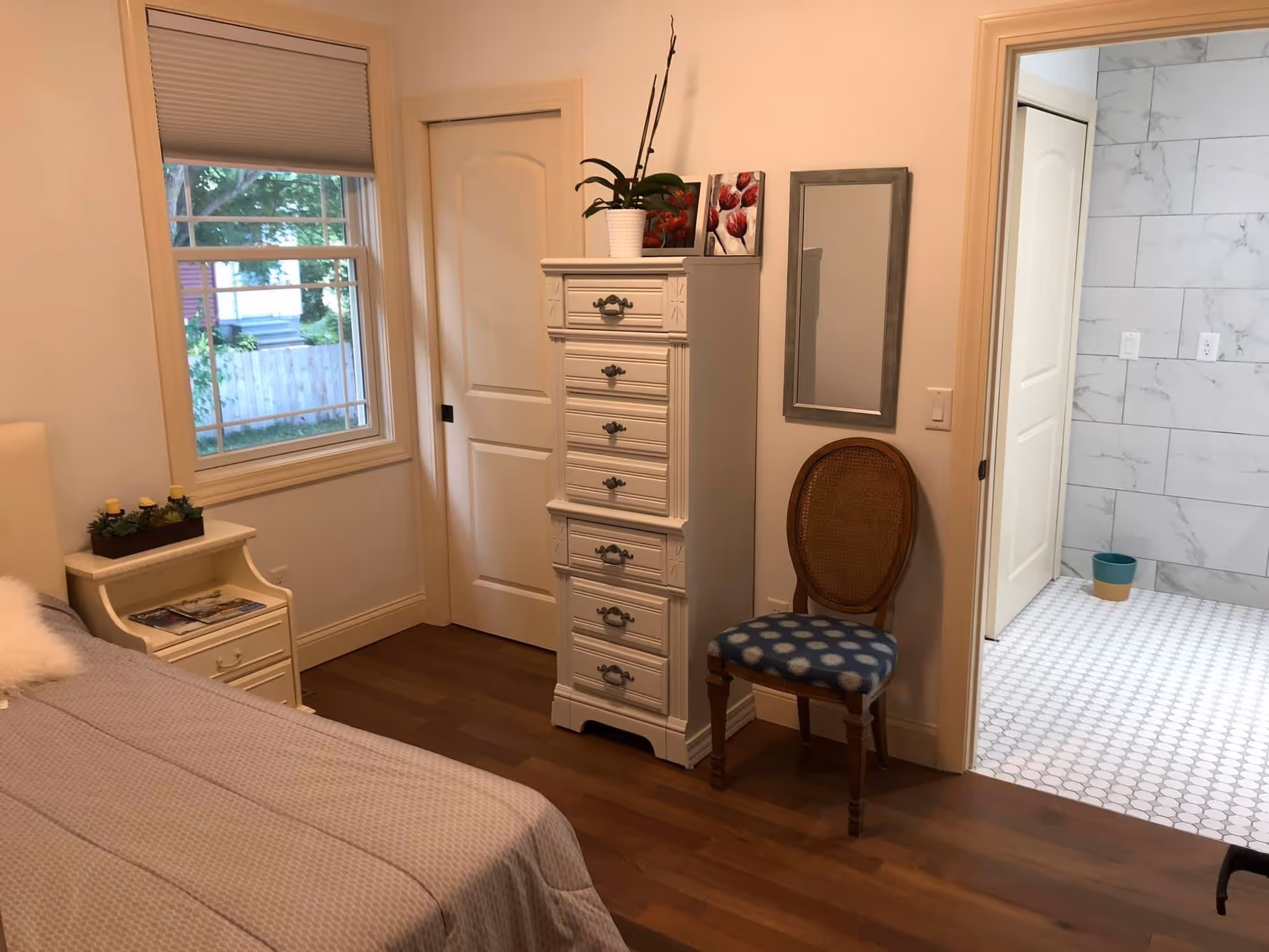 A cozy bedroom with a bed covered in a beige quilt, a small white nightstand with a plant and magazines, a tall white dresser with decorative handles, a wooden chair with a blue patterned cushion, and a wall mirror. A door leads to a bathroom with white hexagonal floor tiles and marble-patterned wall tiles. A window with a beige blind shows a view of a fenced yard with greenery outside.