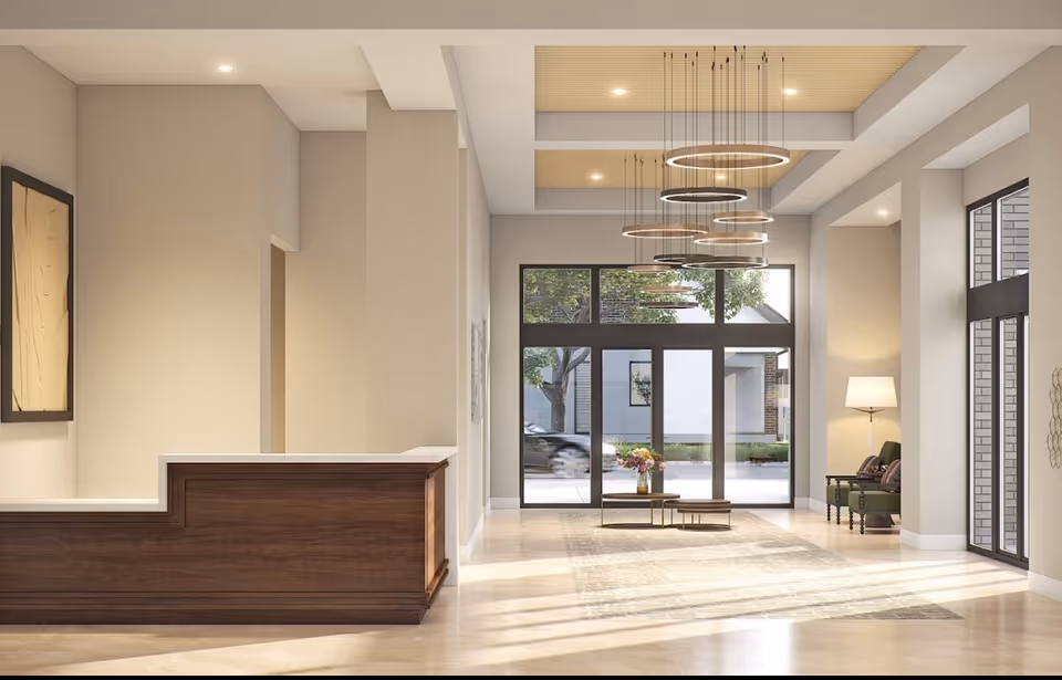A spacious and modern lobby area with a wooden reception desk on the left, large glass doors and windows letting in natural light, a set of circular pendant lights hanging from the ceiling, a small coffee table with flowers, and two green chairs with a floor lamp on the right.