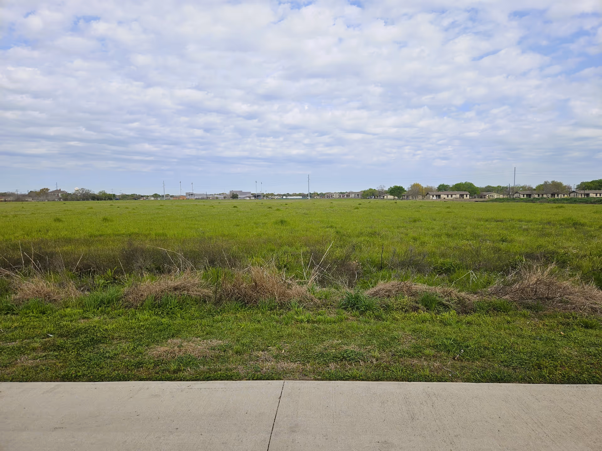 A wide open grassy field with some dry patches and a concrete sidewalk in the foreground. In the distance, there are low buildings and trees under a partly cloudy sky.