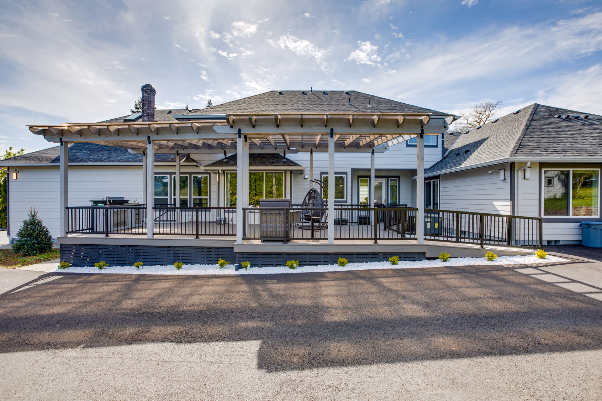 Exterior view of a single-story senior living facility building with a covered porch area featuring outdoor seating and a hanging chair. The building has light-colored siding, a dark shingled roof, and a paved driveway in front. Small shrubs and white decorative stones line the base of the porch.