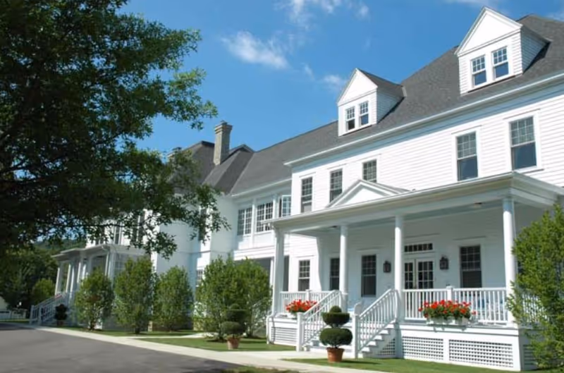 Exterior view of a large white multi-story building with a gray roof, featuring a covered porch with white railings and red flowers in window boxes. There are neatly trimmed bushes and trees along the front, and a clear blue sky above.
