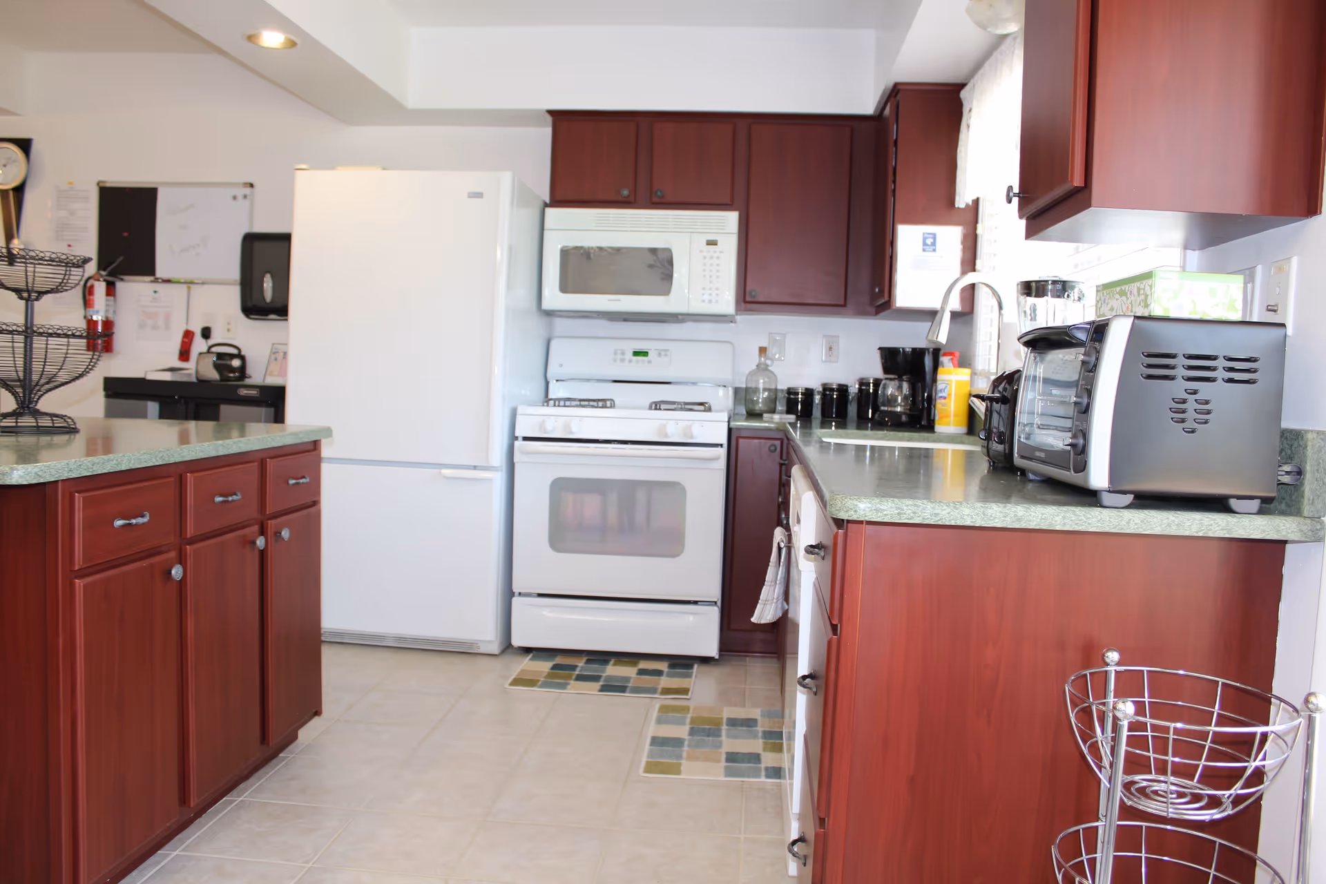 A kitchen with dark wood cabinets, a white refrigerator, white stove with microwave above it, and a countertop with a toaster oven, coffee maker, and other kitchen items. The floor is tiled and there are small patterned rugs in front of the stove and sink.