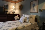 A cozy bedroom with a single bed covered in floral bedding and multiple pillows. There is a wooden dresser with a lamp, a fan, and a TV on top. The walls are decorated with a framed floral picture and a wreath. The room has soft lighting and a calm, homey atmosphere.