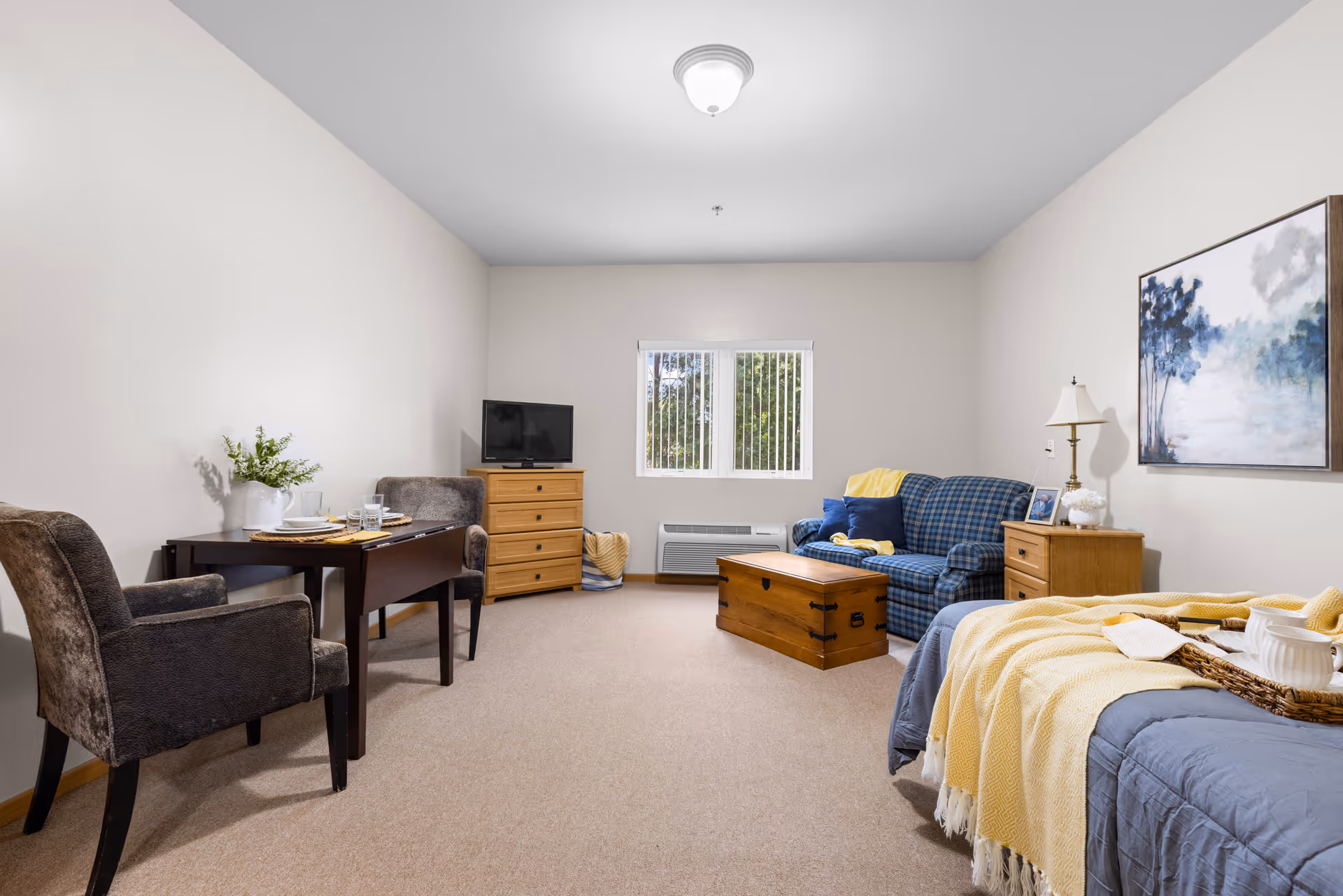 A cozy assisted living room featuring a blue plaid sofa with yellow and blue cushions, a wooden chest coffee table, a wooden dresser with a small TV, a small dining table set with two chairs and tableware, a bed with a yellow blanket and a tray with a teapot and cups, a wooden nightstand with a lamp and picture frame, and a large painting of a misty forest on the wall. A window with vertical blinds lets in natural light.