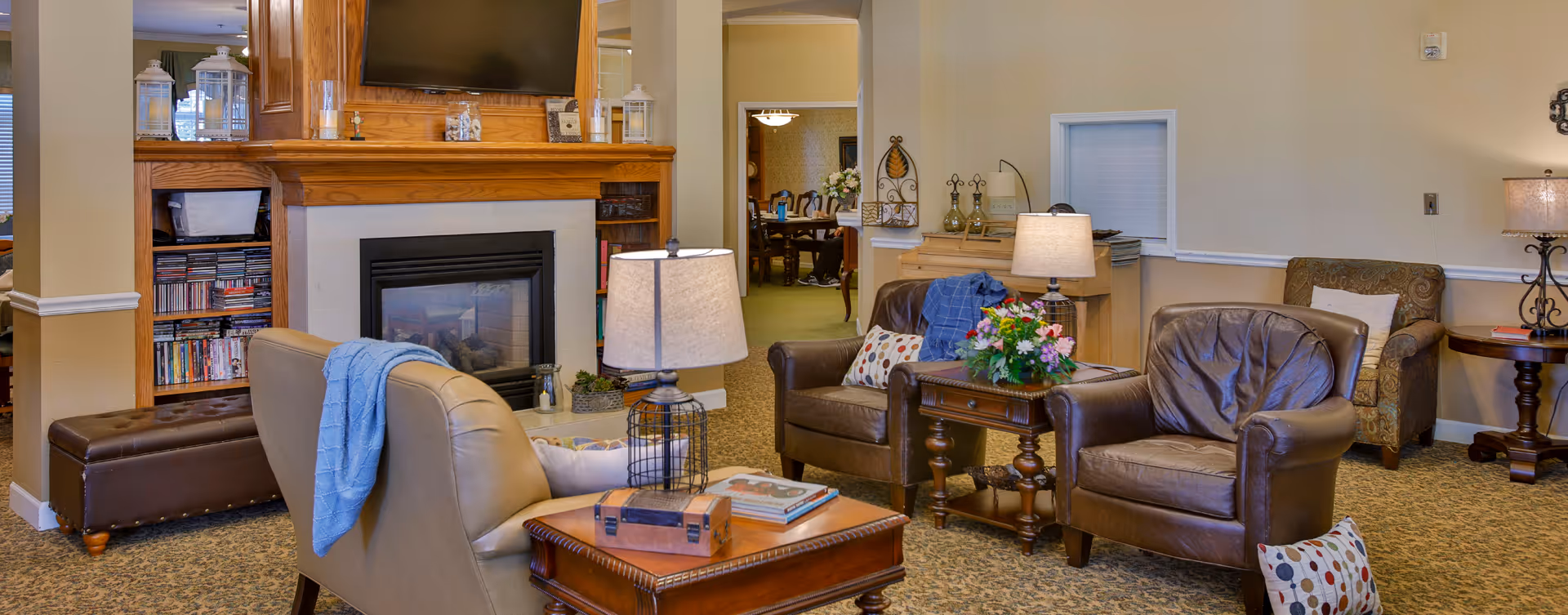 A cozy living room area in a senior living facility featuring a fireplace with a TV mounted above it, surrounded by wooden shelves filled with books and DVDs. The room has comfortable leather and fabric armchairs with decorative pillows and throws, wooden side tables with lamps, and a floral arrangement on one table. In the background, a dining area is visible through an open doorway.