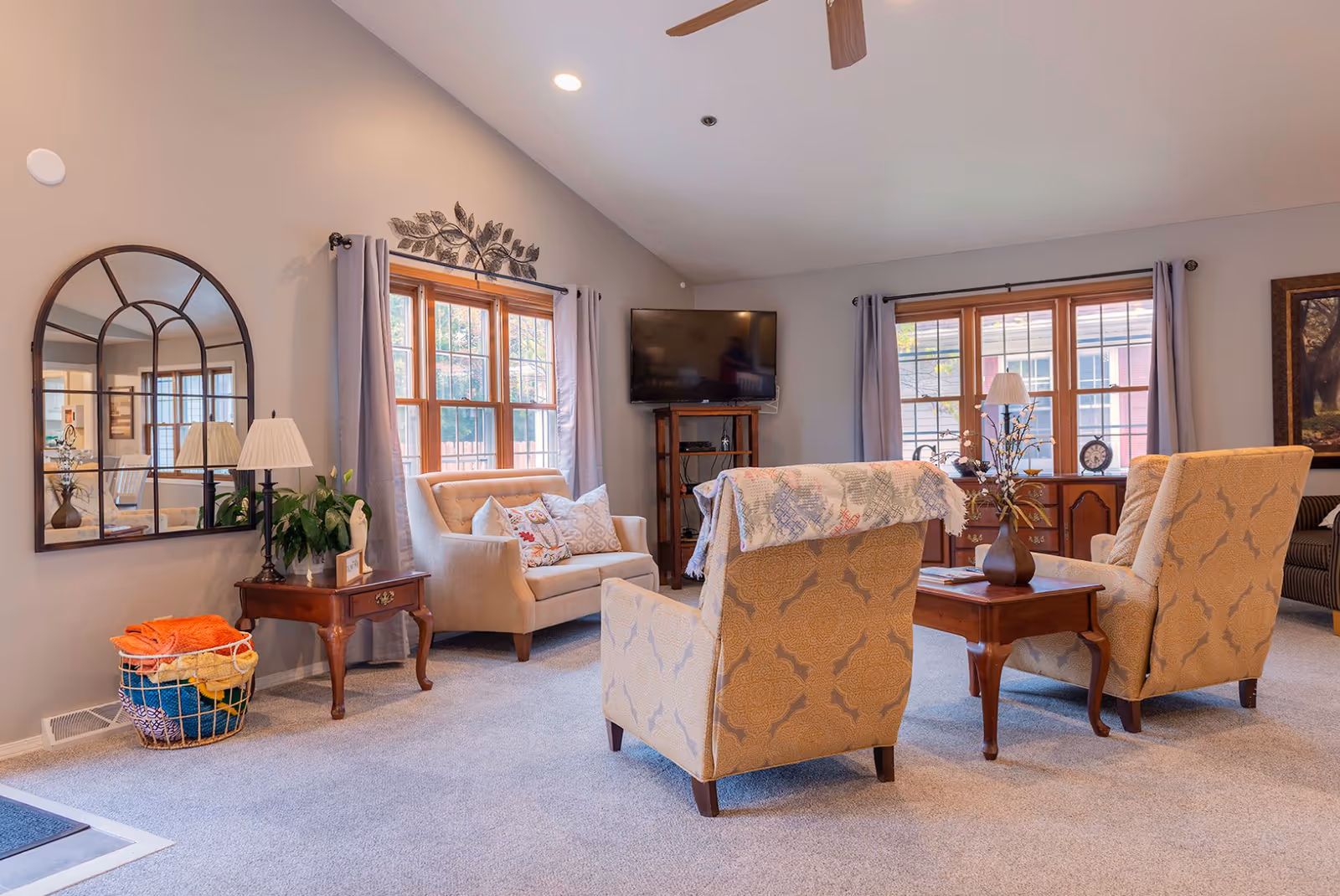 A cozy senior living room with two patterned armchairs, a loveseat with decorative pillows, a wooden coffee table with a vase, and a TV mounted on a stand. The room has large windows with gray curtains, a wall mirror, a side table with a lamp and plant, and a basket with colorful blankets on the floor.