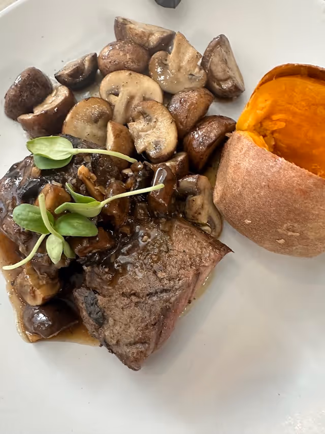 A plated meal featuring a piece of cooked steak topped with sautéed mushrooms and garnished with green sprouts, accompanied by a baked sweet potato with the skin partially peeled back.