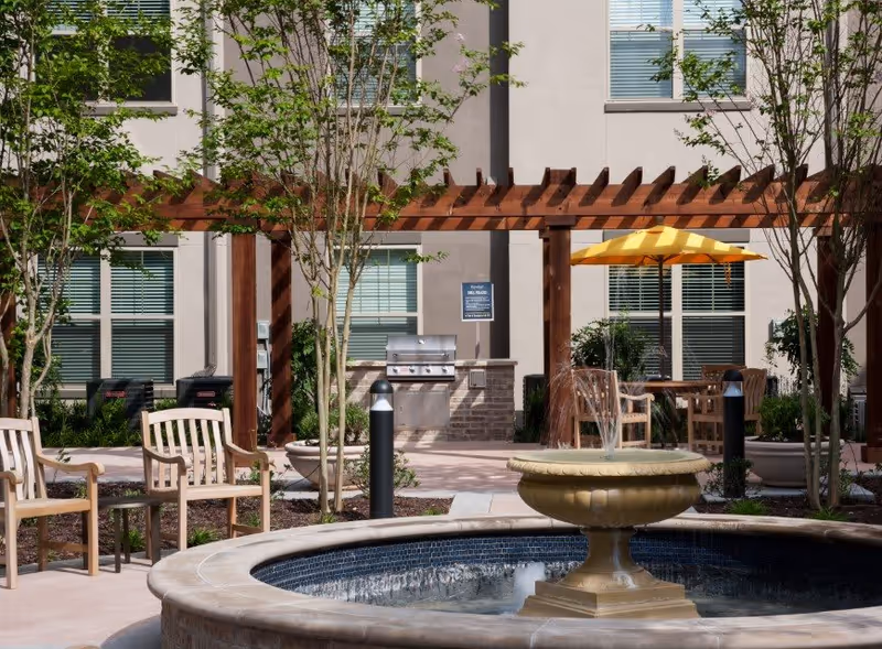 Outdoor courtyard featuring a central fountain, wooden pergola, seating, a yellow umbrella, and a built-in grill in front of the building.