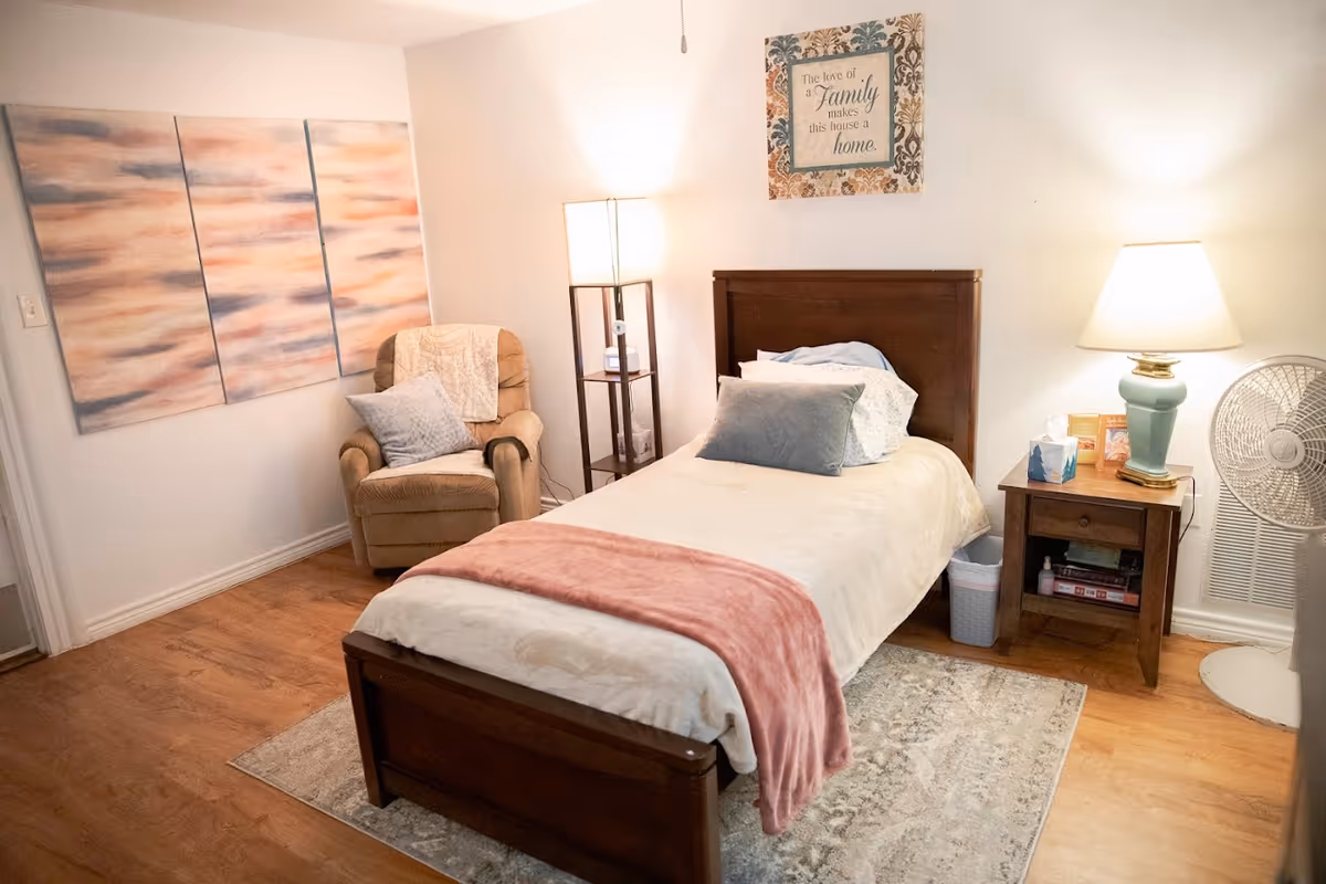 A cozy bedroom with a single wooden bed covered with cream and pink bedding, a beige recliner chair with a pillow and throw blanket, a wooden nightstand with a lamp, tissues, and books, a standing fan, a floor lamp, and wall art including a three-panel abstract painting and a framed quote that reads 'The love of a family makes this house a home.'