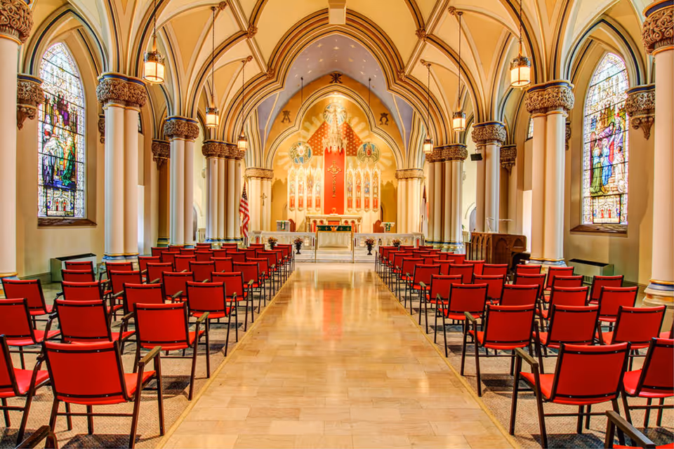 Interior view of a chapel with rows of red chairs leading to an ornate altar, stained glass windows and vaulted arches.