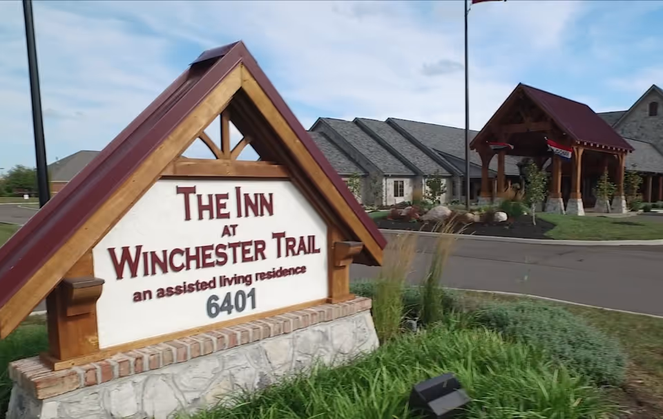Outdoor view of the entrance sign for The Inn at Winchester Trail, an assisted living residence, with the building and covered entryway visible in the background under a partly cloudy sky.