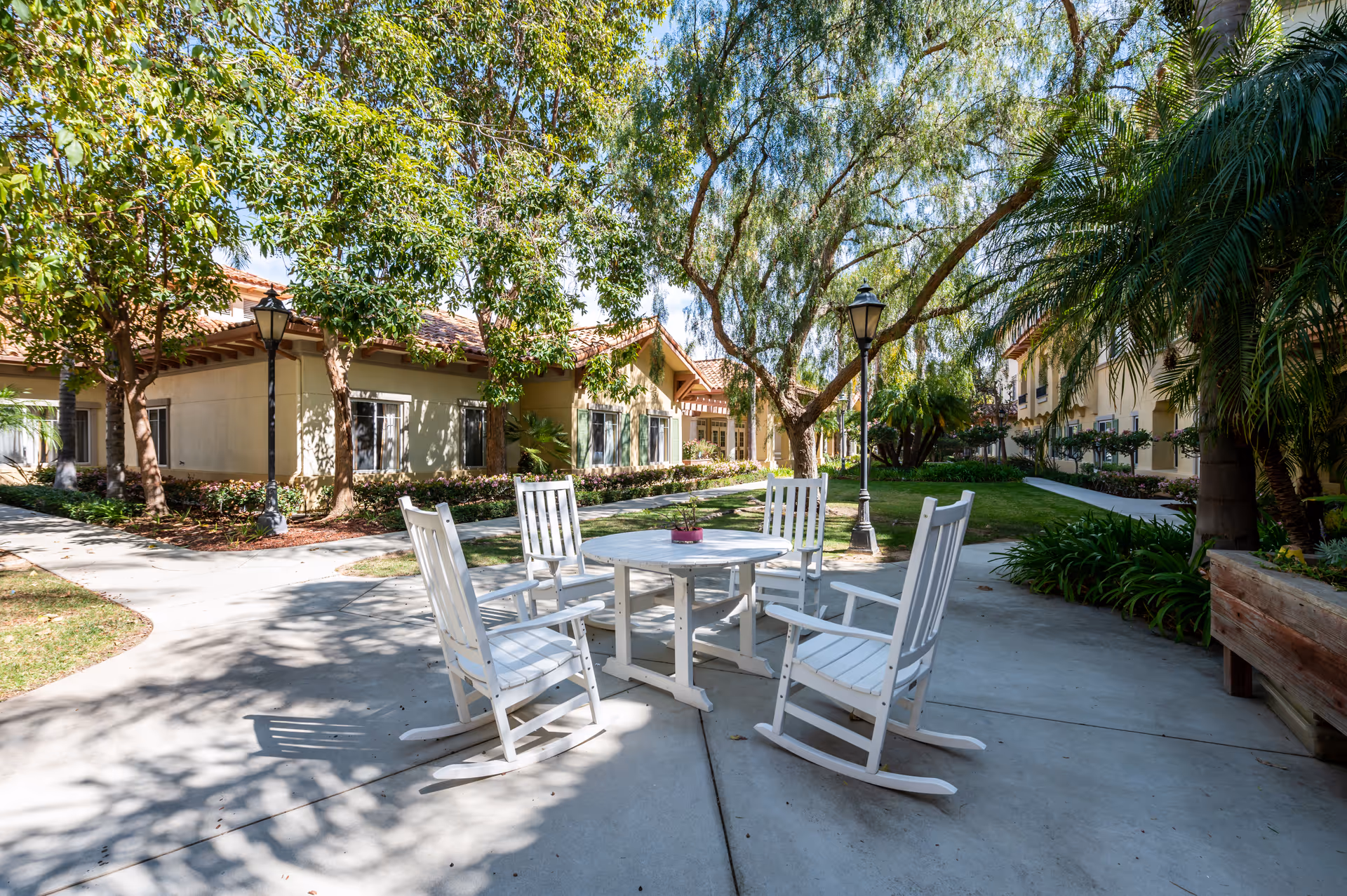 Courtyard with white rocking chairs around a round table beneath trees and lamp posts in front of beige senior living buildings.