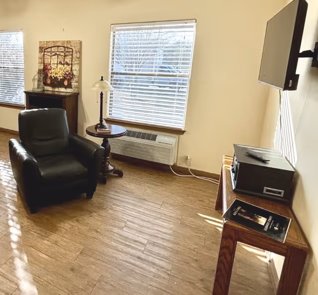 A cozy living room area with a black leather armchair, a round wooden side table with a lamp, two windows with blinds, a wooden cabinet with a decorative flower arrangement, a wall-mounted TV, and a wooden console table holding a stereo system and a magazine.