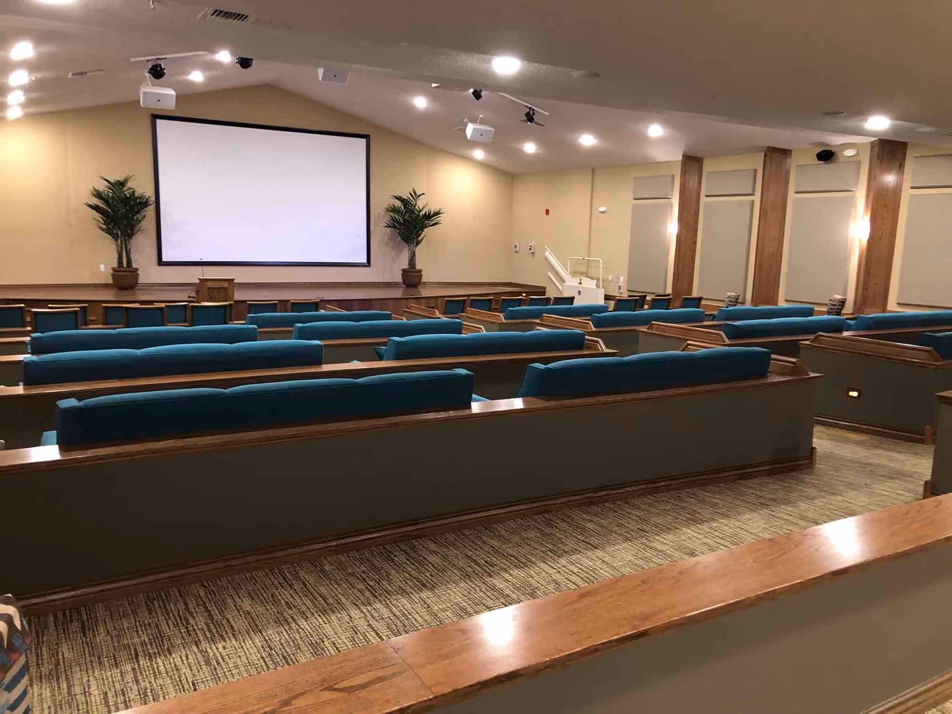 Interior view of a large room with multiple rows of teal cushioned pew-style seating facing a stage with a large blank projection screen and two potted plants on either side. The room has beige walls, carpeted floor, and recessed lighting on the ceiling.