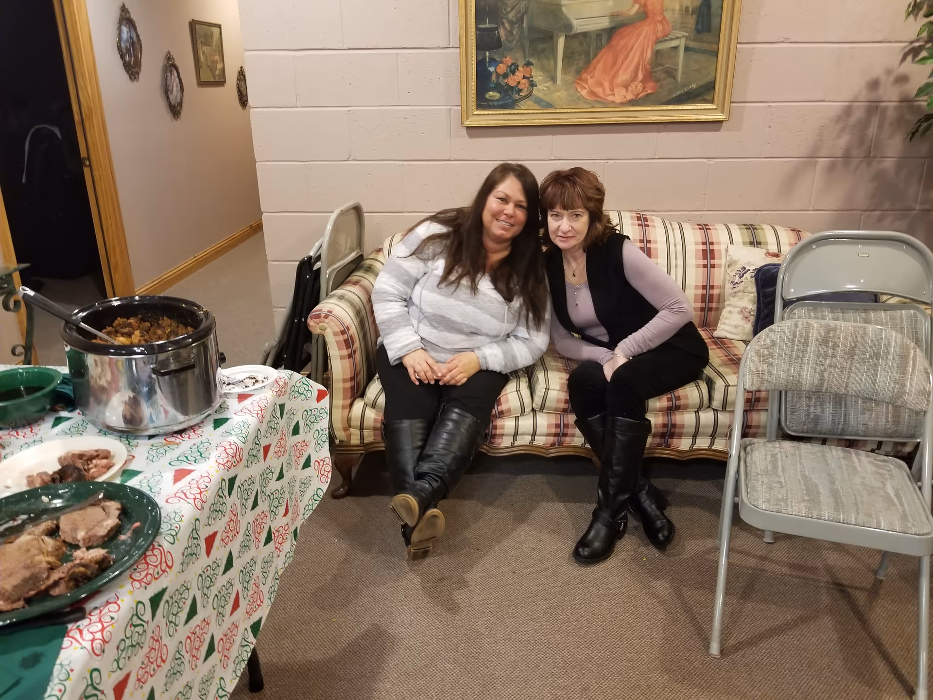 Two women sitting closely together on a plaid patterned couch in a room with beige walls. To the left, there is a table covered with a festive tablecloth holding plates of food and a slow cooker. There are folding chairs next to the couch and framed artwork on the wall behind them.