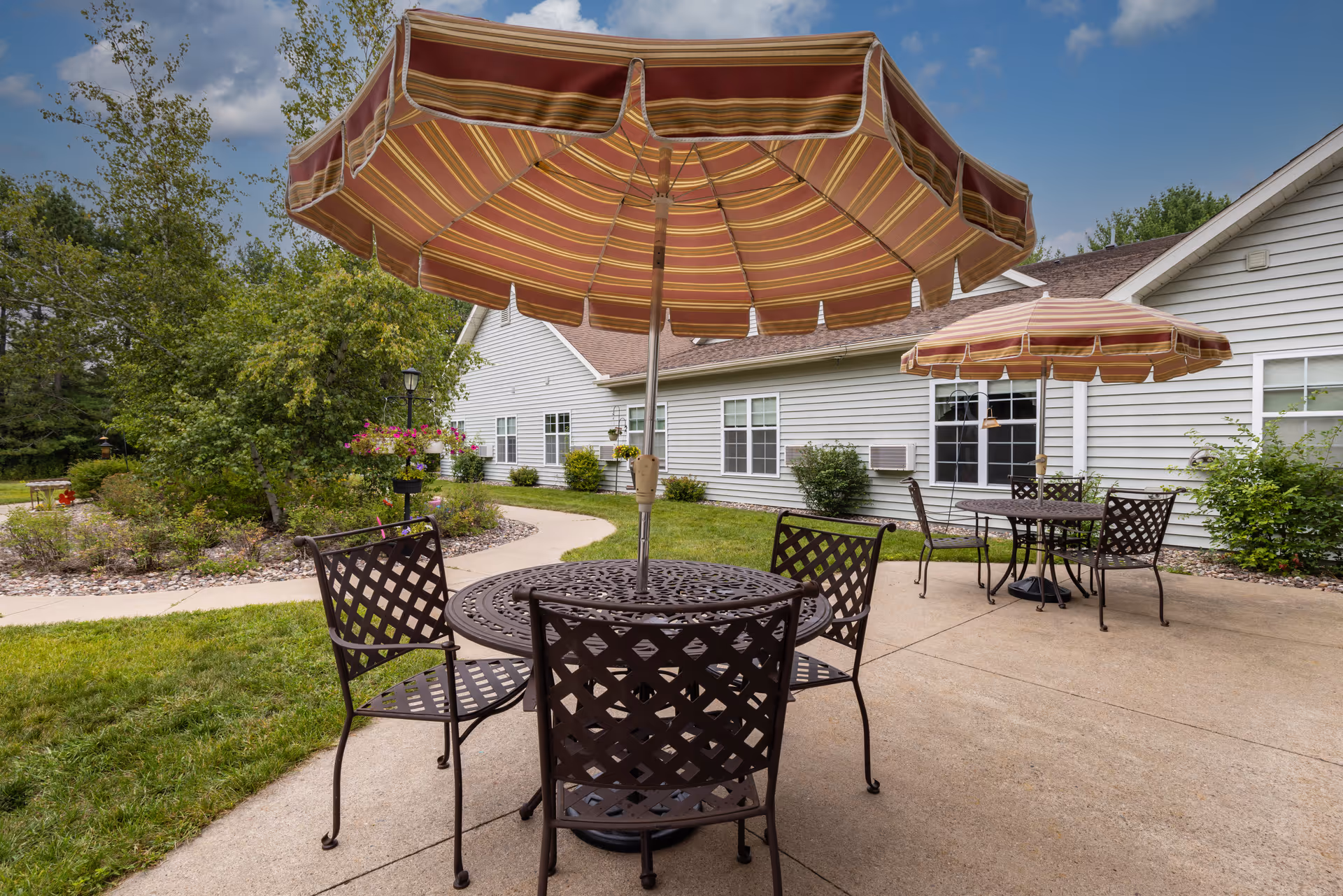 Outdoor patio area at Milestone Senior Living Rhinelander with metal tables and chairs, each table shaded by a large striped umbrella. The patio is adjacent to a light-colored building with windows, surrounded by green grass, bushes, and trees under a partly cloudy sky.