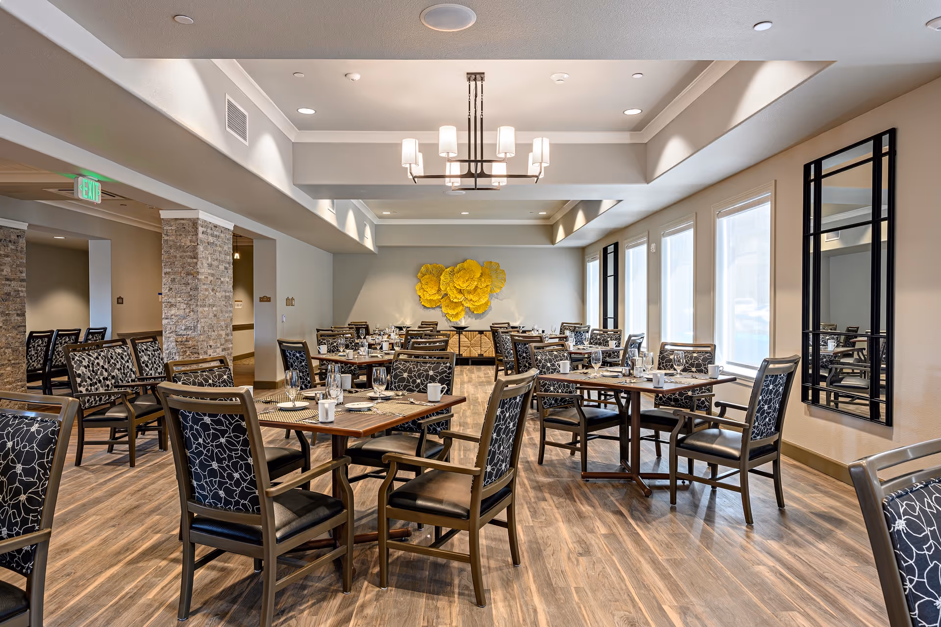 Spacious dining room with multiple set tables and patterned chairs, large windows, and a decorative yellow wall sculpture.