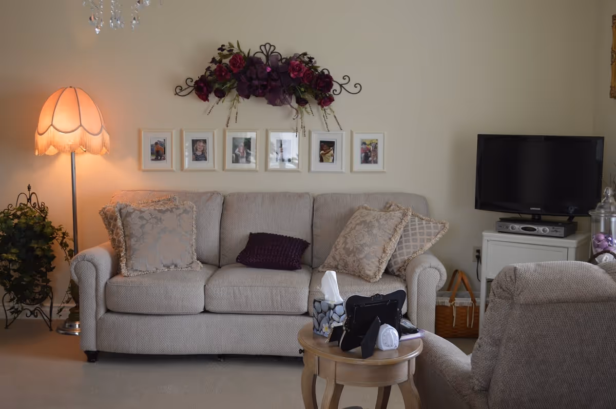 A cozy living room with a beige sofa adorned with decorative pillows, a floor lamp with a fringed shade, a small round wooden side table holding a tissue box, photo frames, and a timer. Above the sofa, there is a wall decoration with purple flowers and six framed photos. To the right, a flat-screen TV sits on a white cabinet with a wicker basket underneath.