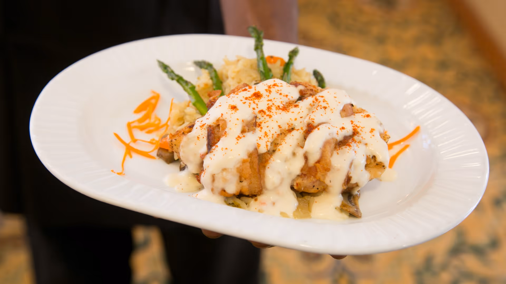 A close-up of a white plate with a serving of food including chicken covered in creamy white sauce, garnished with a sprinkle of red seasoning, asparagus spears, and a side of rice with vegetables. The plate is being held by a person wearing dark clothing.