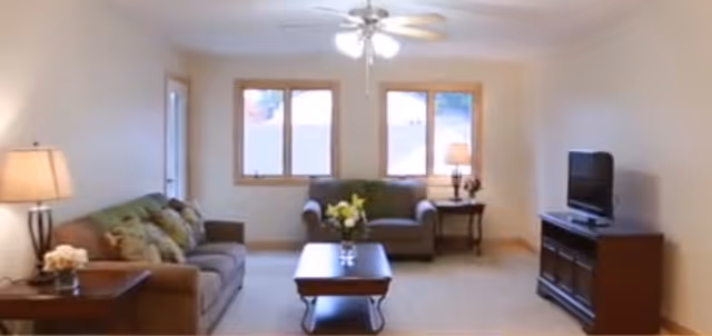 A cozy living room in a senior living community featuring a brown sofa with green and patterned cushions, a matching armchair, a wooden coffee table with a flower vase, a side table with a lamp, and a TV on a wooden stand. The room has beige walls, carpeted floor, and three windows letting in natural light.