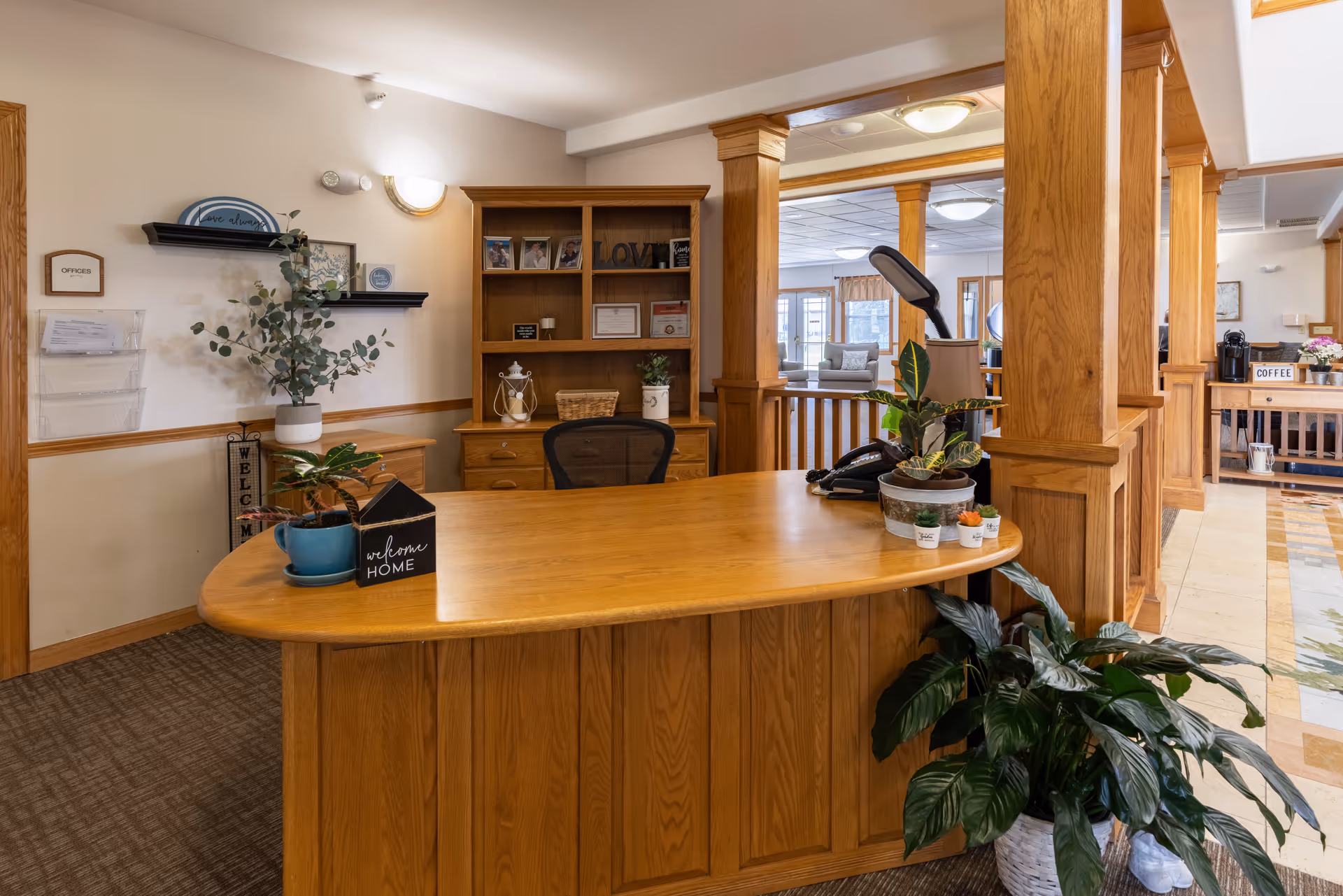Reception desk area in a senior living facility with wooden furniture, potted plants, a phone, and decorative items. Behind the desk is a wooden shelving unit with framed photos and certificates. The area is warmly lit with beige walls and carpeted flooring. In the background, there is a seating area and a coffee station.
