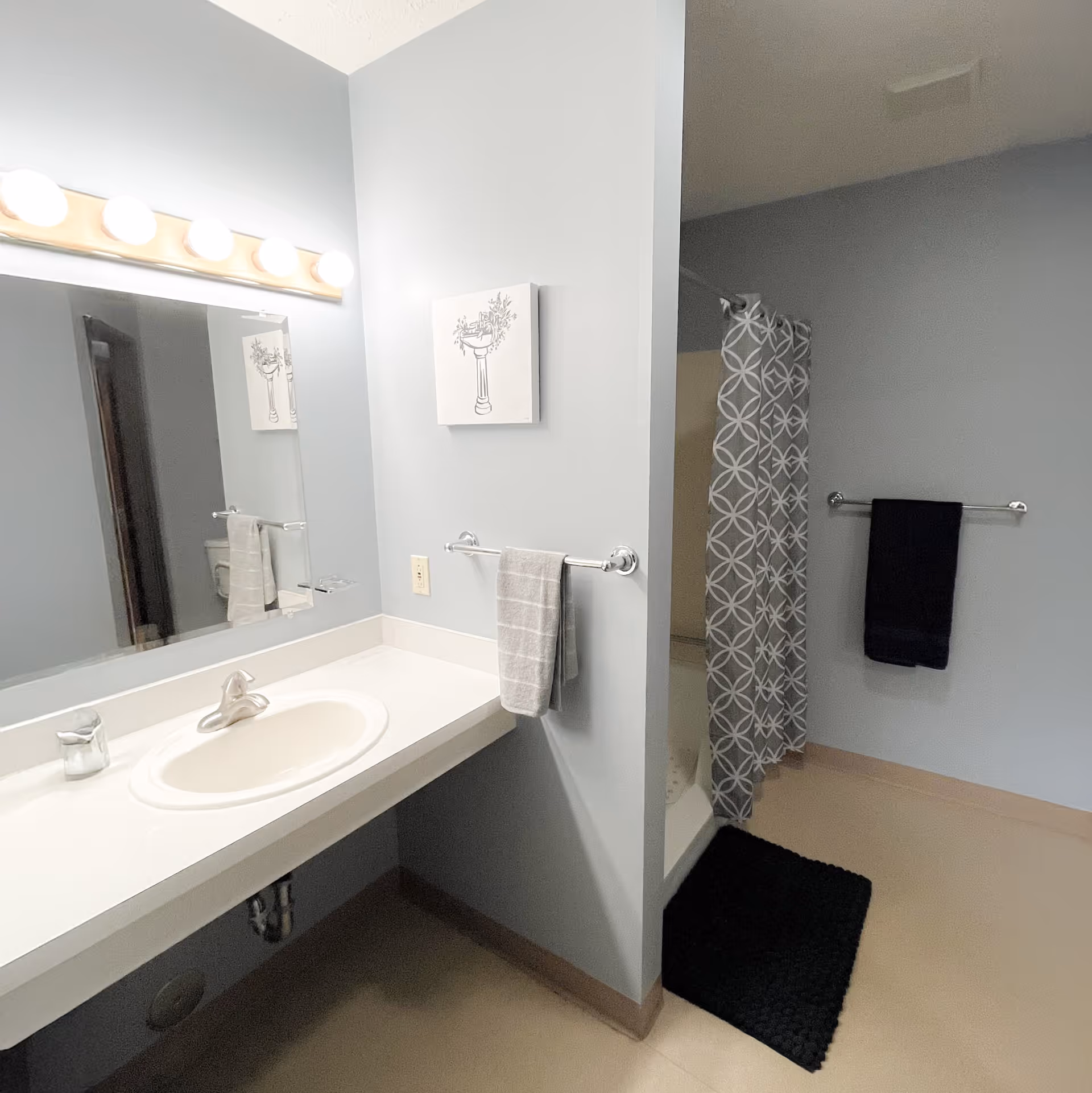 A clean bathroom with a white sink and countertop, a large mirror with five round lights above it, a towel rack holding a gray towel, a shower area with a gray and white patterned curtain, a black towel hanging on a wall-mounted towel rack, and a black bath mat on the floor.