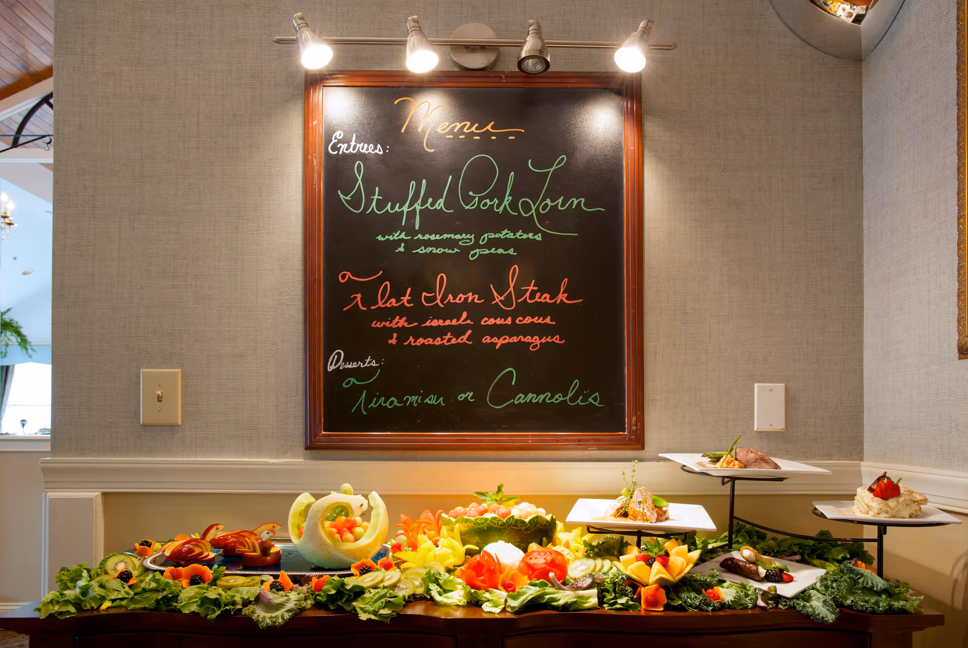 A buffet table with an assortment of decorative fruit carvings and plated dishes, set against a wall with a framed chalkboard menu listing entrees and desserts. The menu includes Stuffed Pork Loin, Flat Iron Steak, and Tiramisu or Cannolis for dessert. The table is garnished with leafy greens and colorful fruits.