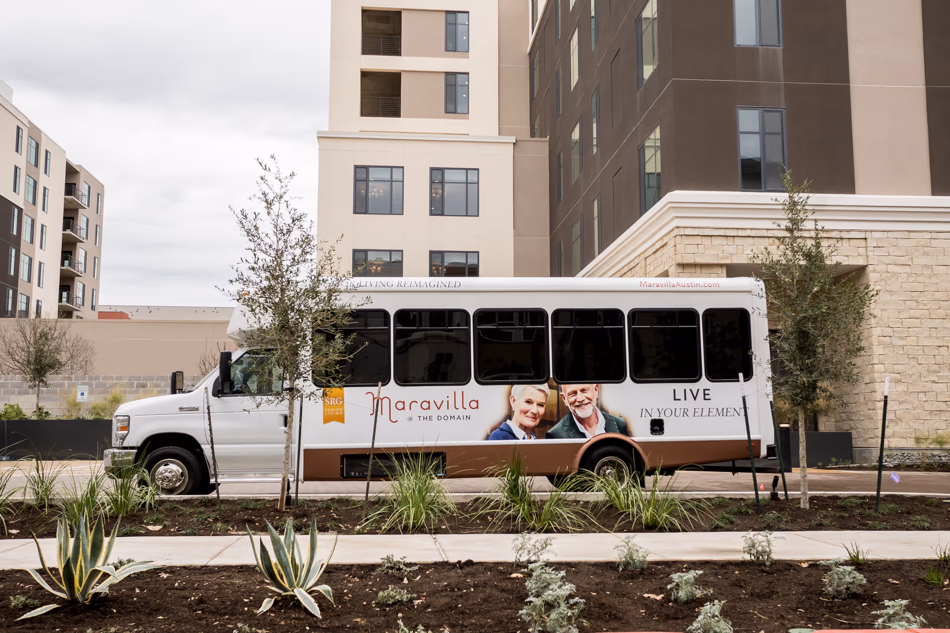 A white shuttle bus parked in front of a multi-story building with beige and brown exterior walls. The bus has signage for Maravilla at The Domain, featuring a logo, images of an elderly man and woman, and the slogan 'LIVE IN YOUR ELEMENT'. There are small trees and plants in the foreground along a sidewalk.