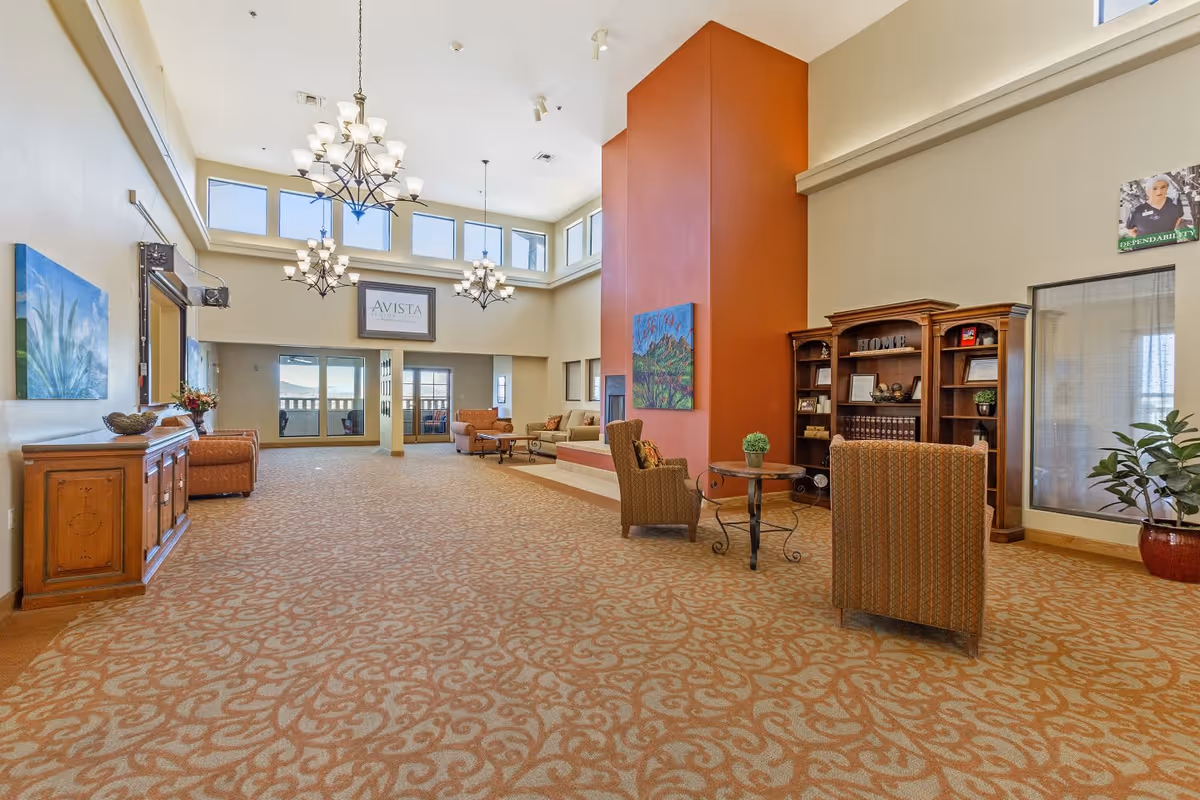Spacious senior living facility common area with high ceilings, large windows near the ceiling, and multiple chandeliers. The room features patterned carpet, comfortable seating including armchairs and sofas, a tall orange accent wall with a painting, a wooden bookshelf, and decorative plants. There is a sign on the far wall that reads 'AVISTA'.