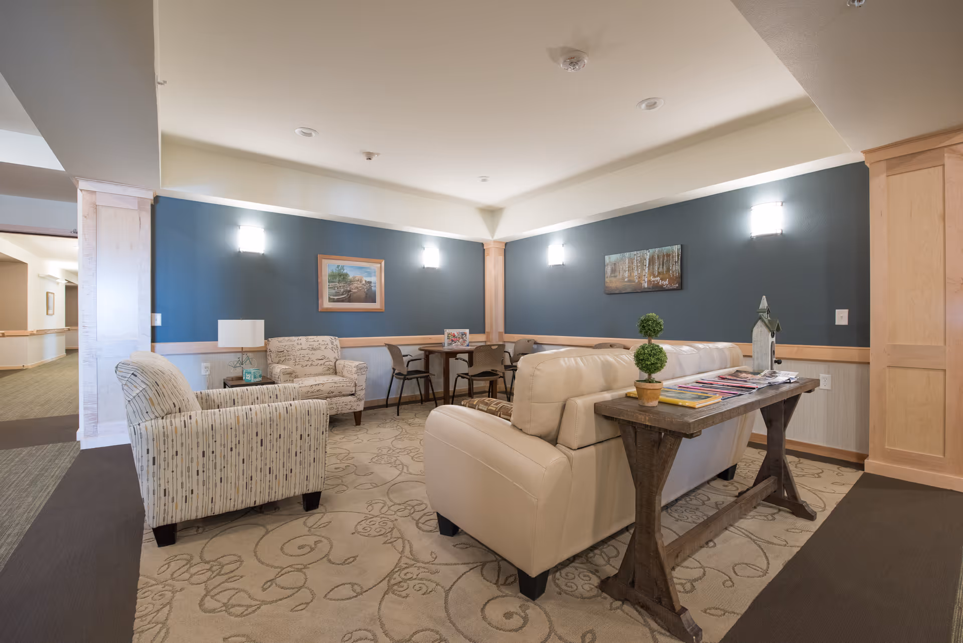 A cozy sitting area in a senior living facility with a beige leather sofa, two patterned armchairs, a wooden table with magazines and small potted plants, and a small dining table with chairs against a blue accent wall adorned with framed artwork and wall sconces.