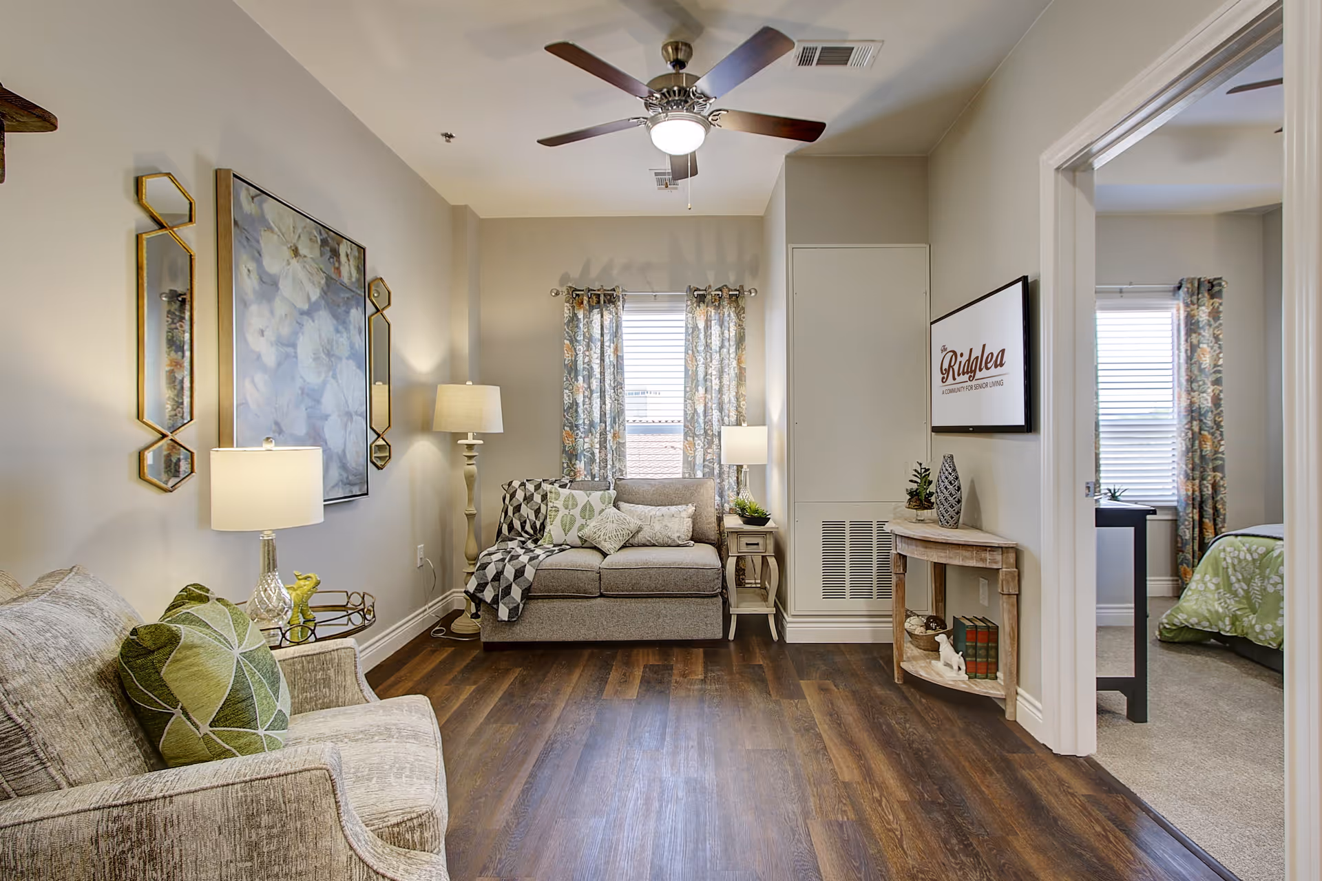 A cozy senior living room with a ceiling fan, two armchairs with green patterned pillows, a small sofa with cushions, two side tables with lamps, floral curtains on the window, and a small wooden console table with decorative items. A doorway leads to a bedroom with a green bedspread and a window with matching curtains. A framed sign on the wall reads 'Ridglea'.