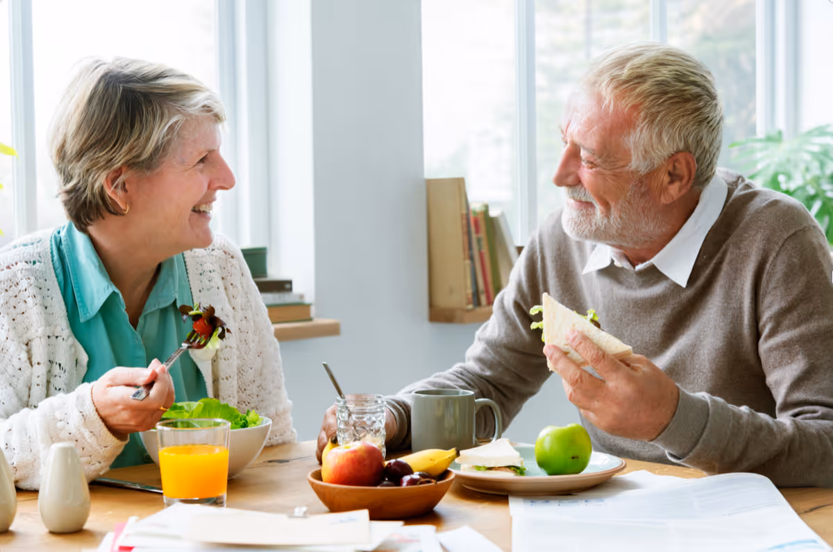An older man and woman sit at a table smiling while eating a meal together in a bright dining area.