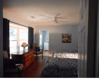 A bedroom with two beds covered in dark patterned bedspreads, a wooden dresser with a vase of flowers on top, two chairs, a ceiling fan, and a doorway leading to a bathroom with a sink and mirror visible.