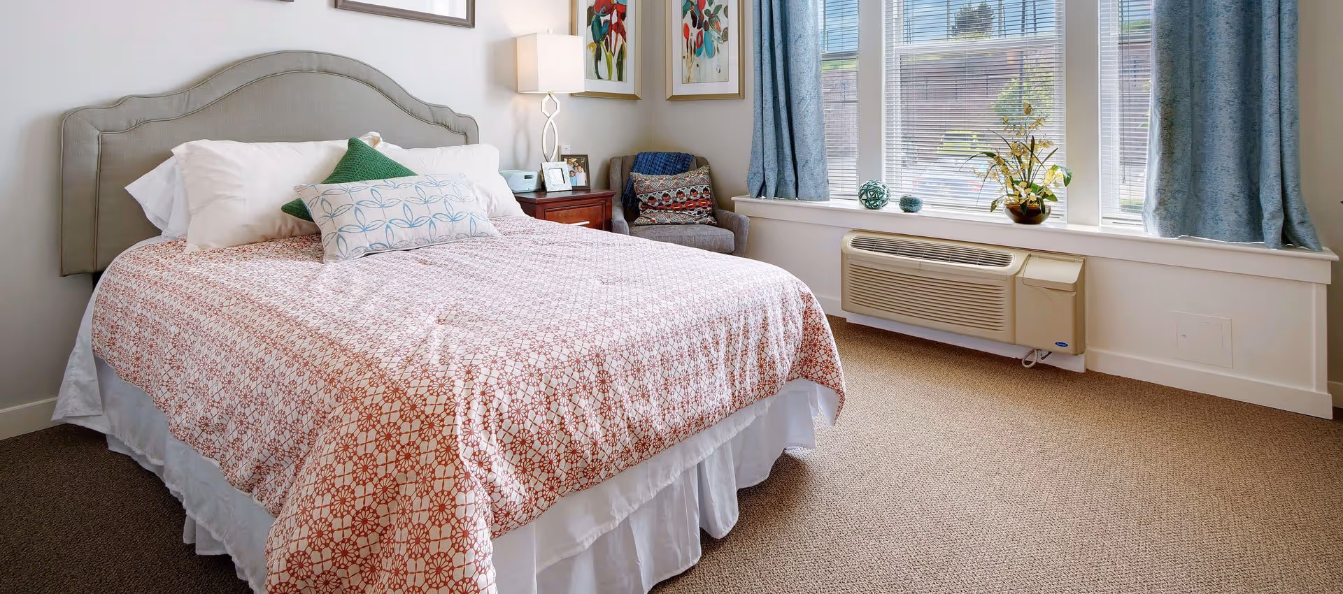 Sunlit bedroom with a made bed, nightstand and armchair beside large windows and a wall air-conditioning unit.