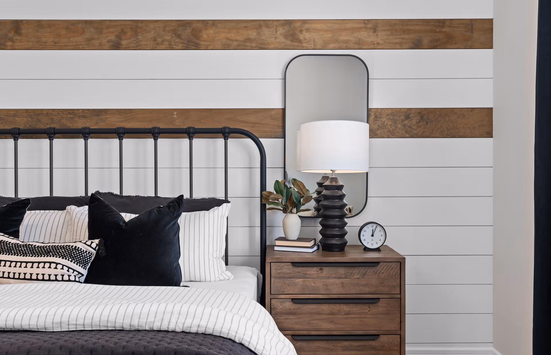 Bedroom featuring a metal bedframe, striped shiplap wall with wood accents, and a wooden nightstand topped with a lamp, mirror, clock, and decor.