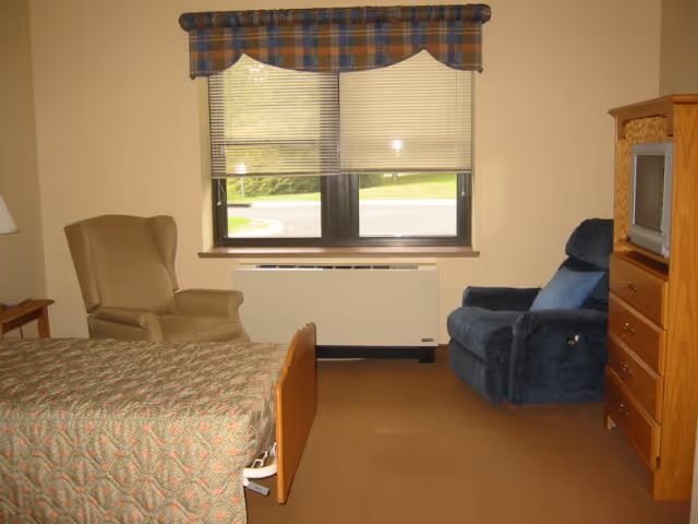 A senior living facility room with a bed covered in a patterned quilt, a beige armchair, a blue recliner, a wooden dresser with a small TV on top, and a window with blinds and a plaid valance. The room has beige walls and carpeted floor.