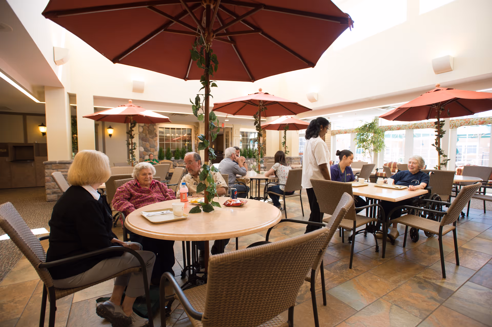 A bright indoor dining area with several elderly people sitting at round tables under large red umbrellas. Two staff members are interacting with the residents. The space has large windows letting in natural light and plants decorating the area.