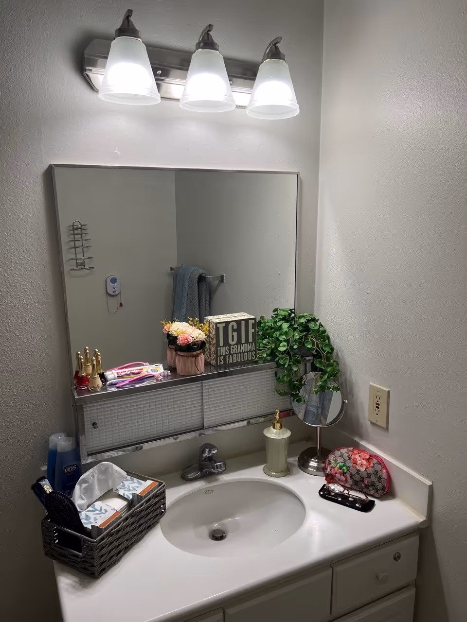 A bathroom sink area with a white countertop and a silver faucet. Above the sink is a rectangular mirror with a three-light fixture. On the countertop, there is a soap dispenser, a small round mirror, a floral-patterned pouch, a pair of glasses, and a basket containing tissues, a hairbrush, and other toiletries. On the shelf below the mirror, there are small decorative items including a sign that reads 'TGIF THIS GRANDMA IS FABULOUS,' a small plant, and some nail polish bottles. A towel hangs on a rack reflected in the mirror.