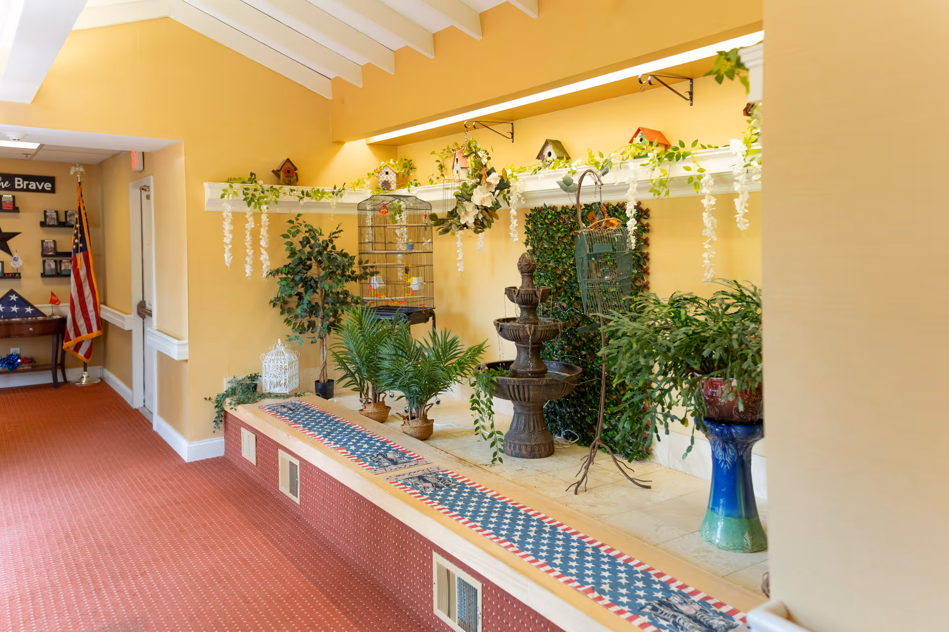 Indoor senior living hallway with decorative plants, birdcages and a tiered fountain beside a raised planter area.