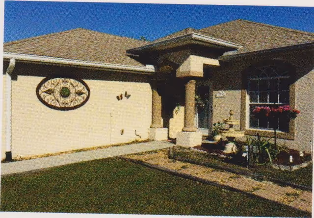 Exterior view of Cobblestone Manor Assisted Living Facility showing a beige building with a shingled roof, a small garden area with plants and flowers, a decorative wall ornament, and a covered entrance supported by two columns under a clear blue sky.