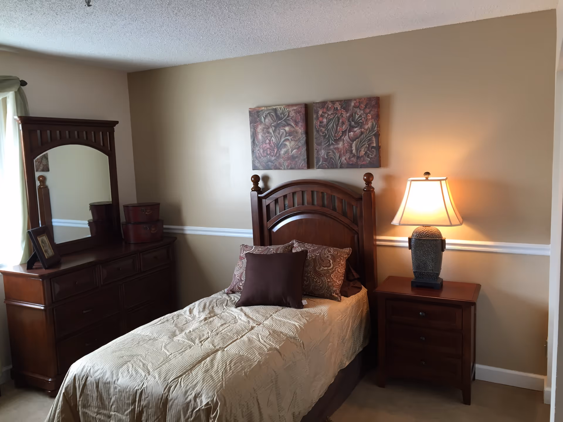 A bedroom with a single bed featuring a wooden headboard, beige bedspread, and decorative pillows. To the right of the bed is a wooden nightstand with a lamp turned on. On the left side of the room is a wooden dresser with a large mirror and two decorative boxes on top. The walls are painted beige with a white chair rail molding, and two framed floral artworks hang above the bed.